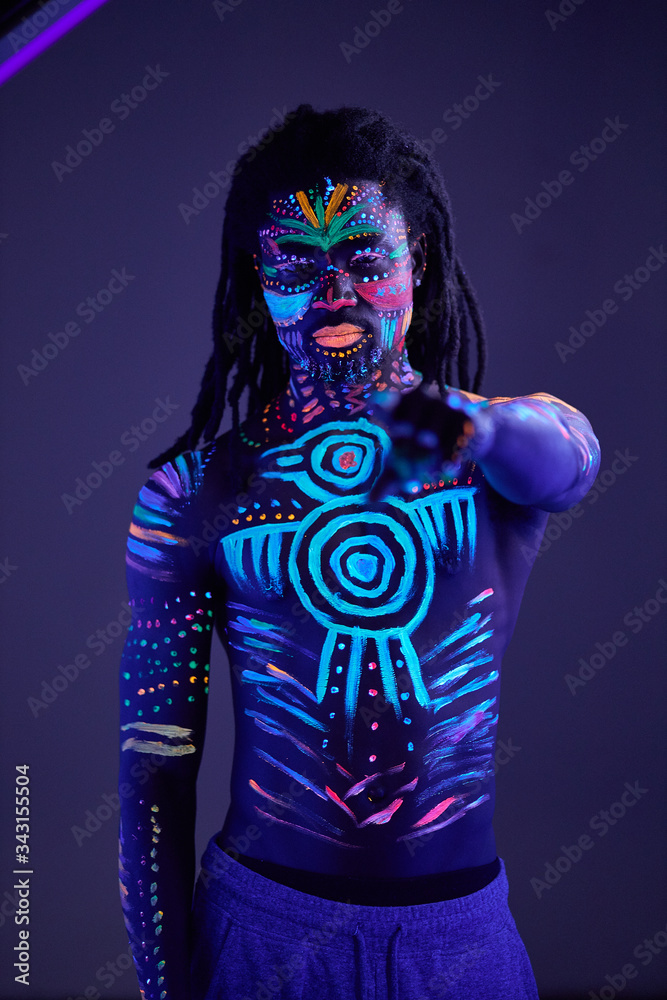 portrait of african male with fluorescent body art glowing in neon lights in dark space. fashion model with naked skin aim his finger at camera portrait of african male with fluorescent body art glowing in neon lights in dark space. fashion model with naked skin aim his finger at camera