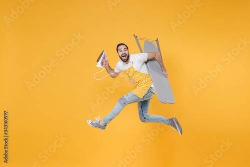 Side view of excited young man househusband in apron rubber gloves hold iron board for ironing while doing housework isolated on yellow background studio. Housekeeping concept. Jumping looking camera.