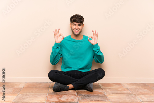 Young handsome man sitting on the floor showing an ok sign with fingers