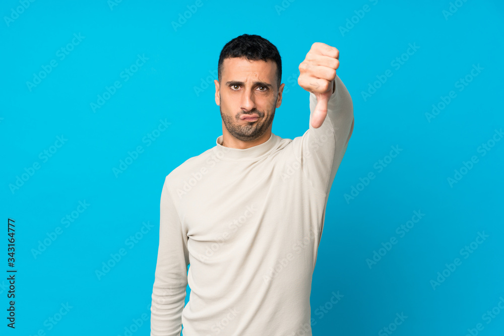 Young man over isolated blue background showing thumb down with negative expression Young man over isolated blue background showing thumb down with negative expression