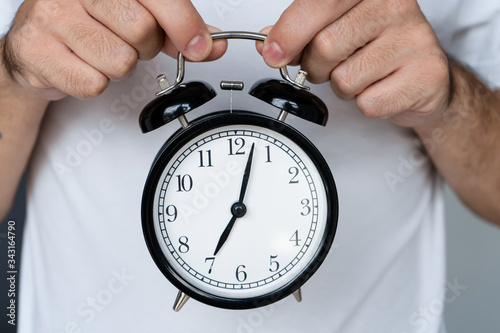 A guy in a white T-shirt is holding a stylish black alarm clock with a bell. On the alarm clock, the beginning of the eighth. Time to get up.