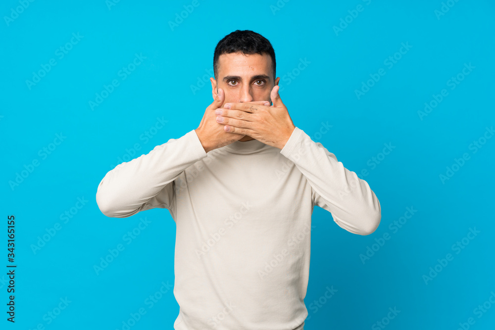 Young man over isolated blue background covering mouth with hands Young man over isolated blue background covering mouth with hands