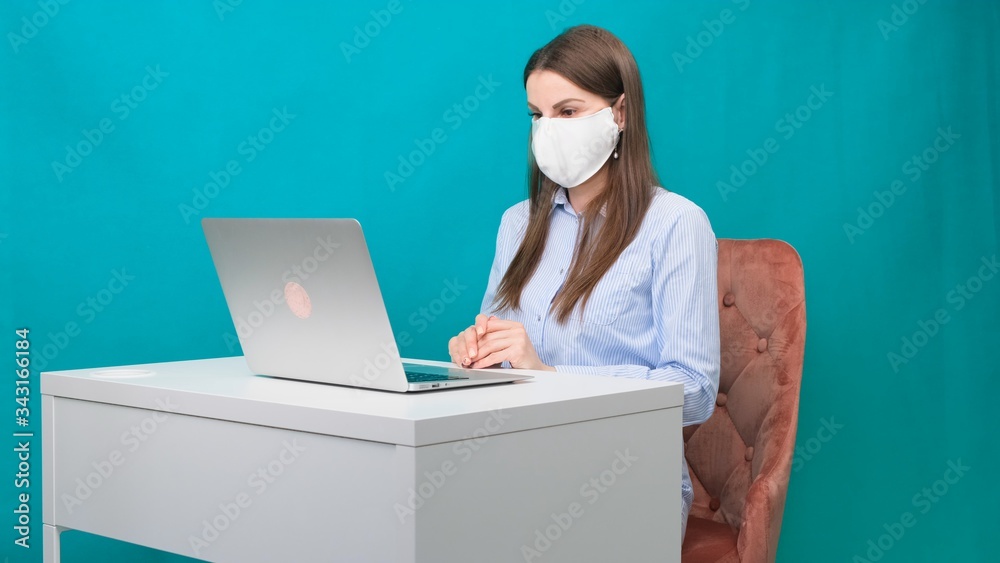 Female in protective mask works on laptop at workplace or at home during a pandemic. The concept of work during quarantine and self-isolation Female in protective mask works on laptop at workplace or at home during a pandemic. The concept of work during quarantine and self-isolation