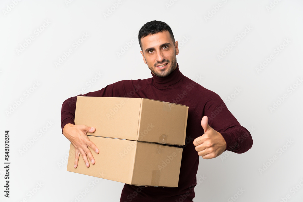 Young man over isolated white background holding a box to move it to another site with thumb up Young man over isolated white background holding a box to move it to another site with thumb up