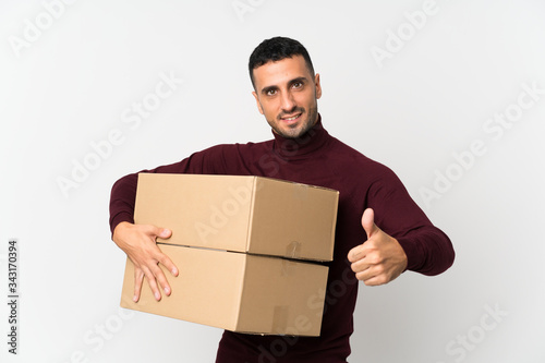 Young man over isolated white background holding a box to move it to another site with thumb up