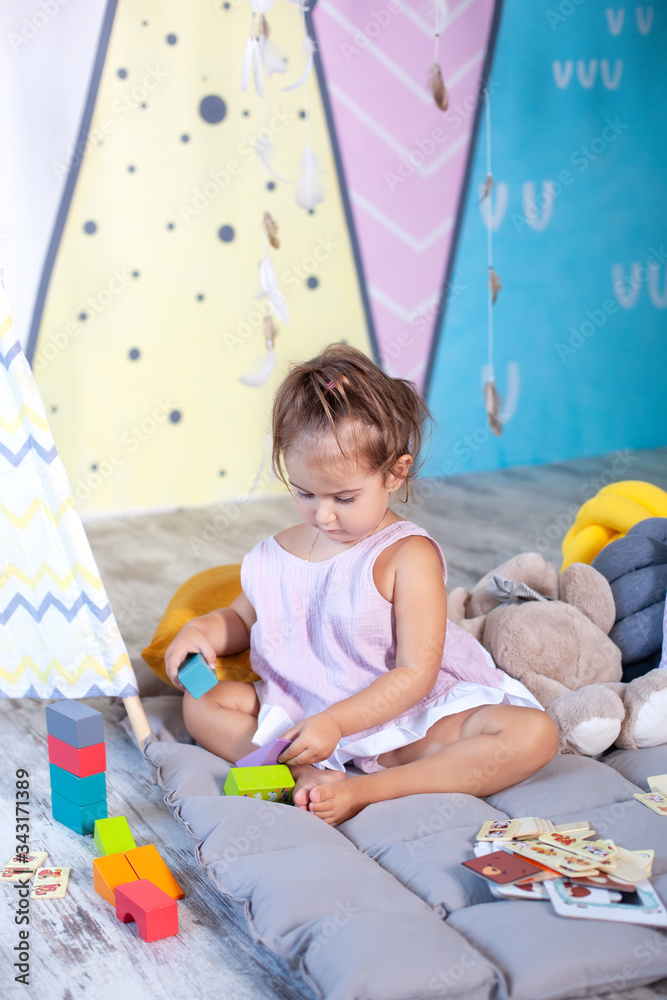 Fototapeta premium Little girl plays wigwam with colorful toy bricks and builds tower. Happy baby plays in tent in children's room with educational toys. baby plays in kindergarten. childhood concept, child development 