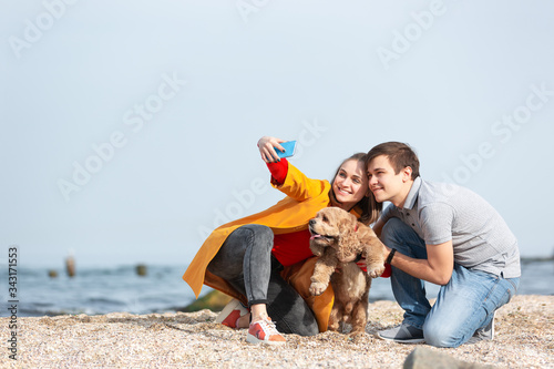 Young couple playing with a dog on a sea coast and making selfie with a mobile phone.
