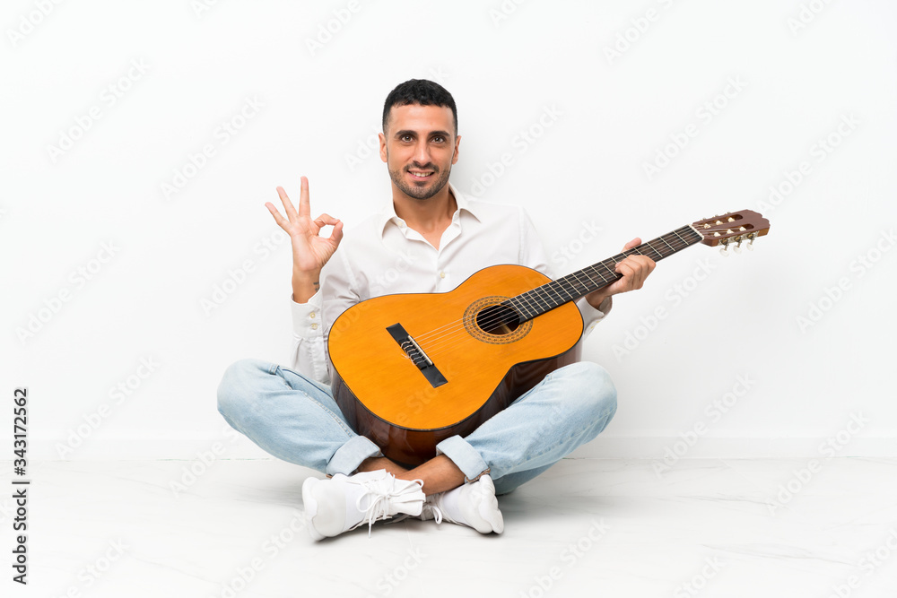 Young man sitting on the floor with guitar showing ok sign with fingers
