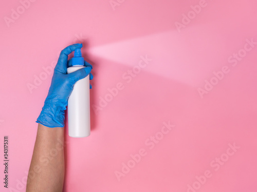 Coronavirus protection. Hand in blue disposable glove overspraying surface disinfectant or hand sanitizer in white spray bottle on pink background. Hygiene measures to prevent Covid-19 pandemic