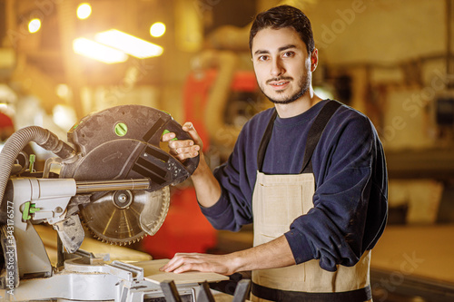 Εκτύπωση καμβά portrait of confident caucasian woodworker at work place, man in uniform enjoy his job, stand behind circular saw machine and look at camera