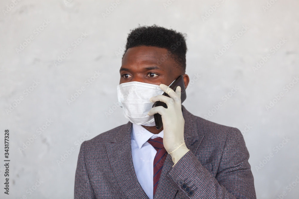 COVID-19. Handsome African black man in a suit is using medical mask and holding his mobile phone with a hand in gloves. Measures to prevent coronavirus infection and the development of a pandemic COVID-19. Handsome African black man in a suit is using medical mask and holding his mobile phone with a hand in gloves. Measures to prevent coronavirus infection and the development of a pandemic