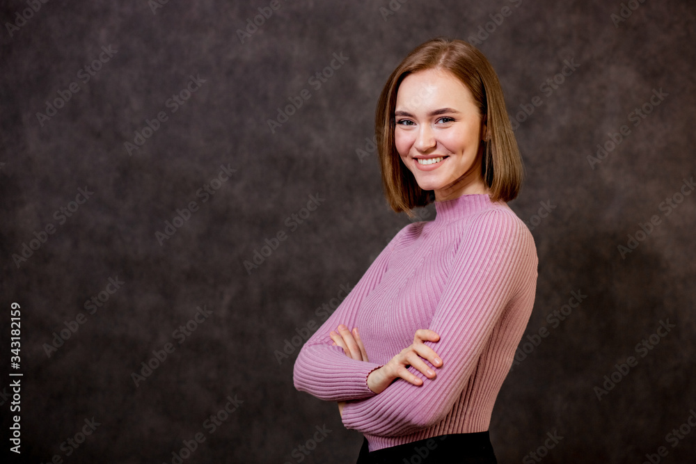 A young beautiful girl crossed her arms over her chest, stands sideways and looks at the camera with a smile, background brown