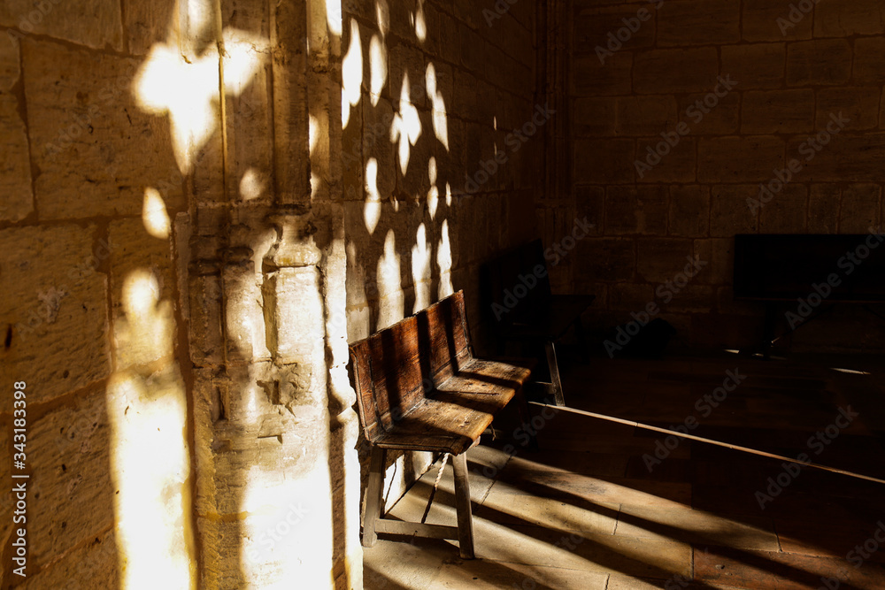 A wooden bench stands in the corner of the annex to the old stone church and the bright day sun with its rays through the openwork lattice draws elaborate patterns on the bench and the wall above it