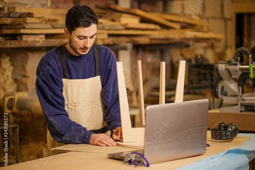 young furniture maker skillfully make a wooden chair on a workbench and use laptop while working alone in his large woodworking shop, handicraft and carpentry concept photo