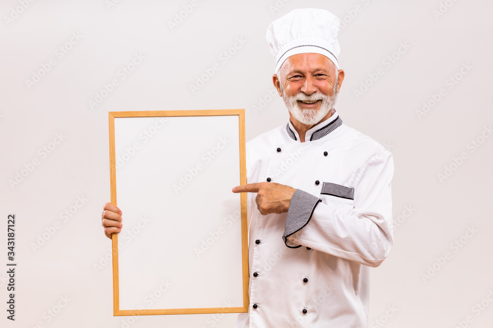 Portrait of senior chef pointing at  whiteboard on gray background.