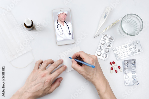Top view - a table with pills, thermometers and water. Patient's hand writes a prescription pen while having a video chat with a doctor on a smartphone. Medicine, technology and healthcare concept.