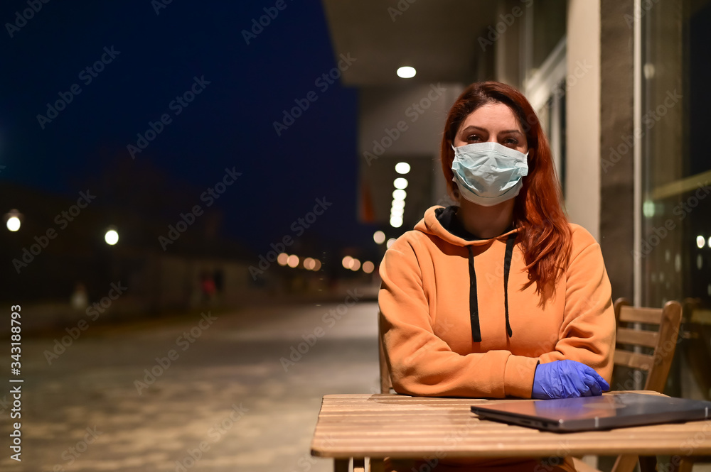 A woman in a medical mask and gloves walks late at night alone. The girl is sitting at a wooden table with a laptop in quarantine. Coronavirus.
