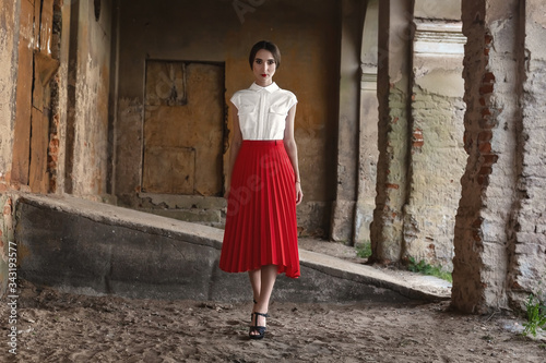 Fashion Model Girl with brown hair against the background of an old, abandoned building in a stylish red skirt and shirt
