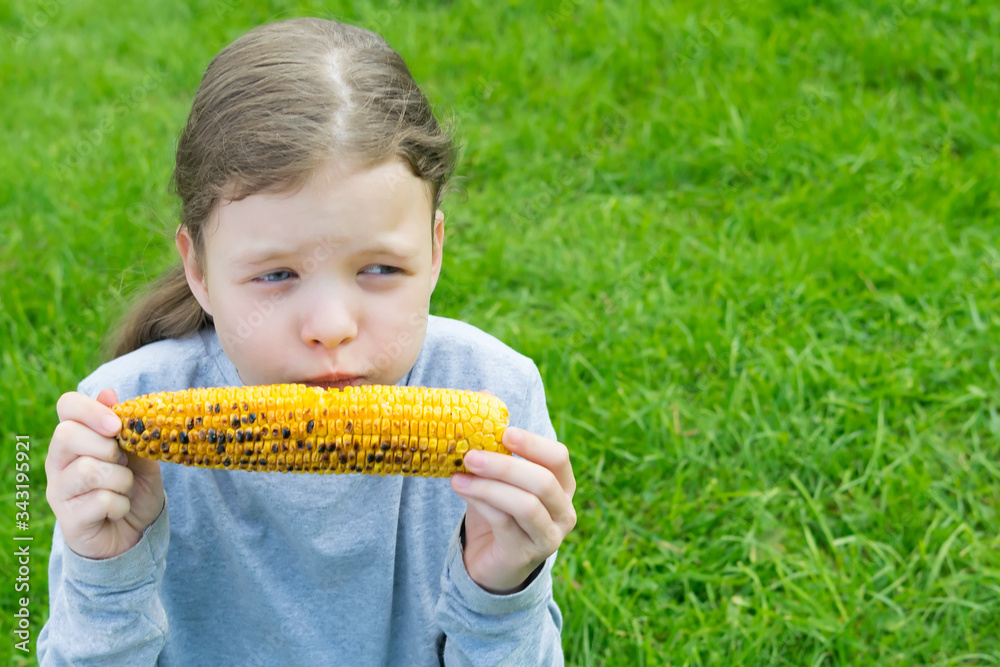 little girl holding a fried yellow corn cob in her hands and wants to eat it, sitting on the green grass little girl holding a fried yellow corn cob in her hands and wants to eat it, sitting on the green grass