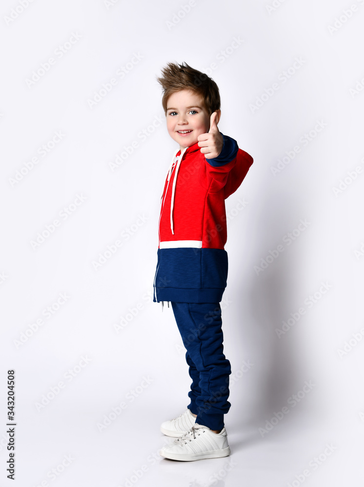 Little blond son in blue and red tracksuit, sneakers. Smiling, showing thumb up, posing sideways isolated on white background Little blond son in blue and red tracksuit, sneakers. Smiling, showing thumb up, posing sideways isolated on white background
