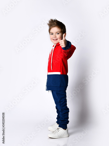 Little blond son in blue and red tracksuit, sneakers. Smiling, showing thumb up, posing sideways isolated on white background photo