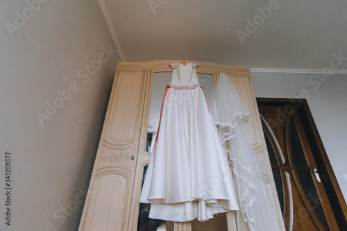 Wedding white long dress of the bride hangs on a hanger, on a wooden wardrobe with a mirror in the hotel room. Photography, concept.