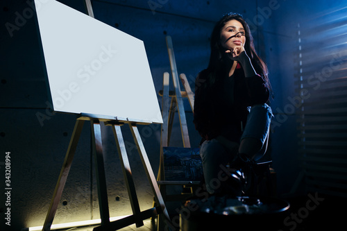 Blank Mock Up Canvas. Beautiful brunette girl artist sitting in a dark Studio near the easel with painted watercolor abstract painting.