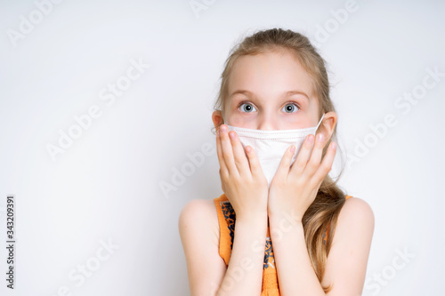 Blonde little girl in orange dress and medical mask, posing isolated on white. She is looking scared, touching her face. Coronavirus. Quarantine. Worldwide pandemic COVID-19. Close up, copy space