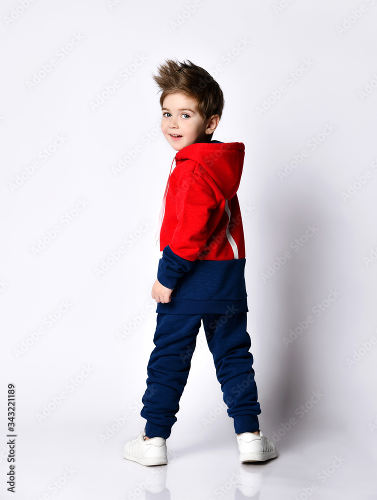 Little blond boy in blue and red tracksuit, sneakers, sunglasses. Smiling turning back, showing thumb up, posing isolated on white Little blond boy in blue and red tracksuit, sneakers, sunglasses. Smiling turning back, showing thumb up, posing isolated on white