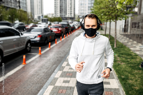 Keep yourself in shape during quarantine. Athletic young man is jogging in a protective mask among high-rise buildings. Healthy lifestyle concept
