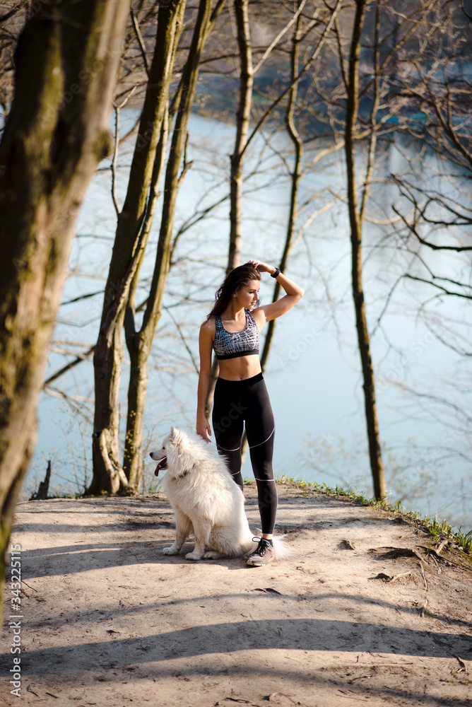 Full height portrait of a young sports girl with the dog on a sunny spring morning in the park. A woman stands next to her dog on a hill near the lake Full height portrait of a young sports girl with the dog on a sunny spring morning in the park. A woman stands next to her dog on a hill near the lake