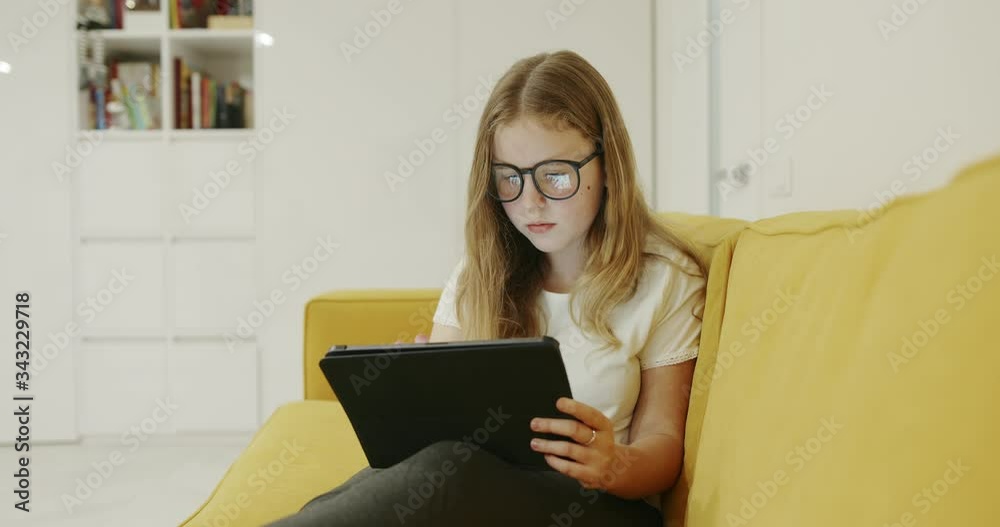 Young blond girl wearing glasses sitting on a comfortable sofa at home using a tablet-pc during the Covid-19 pandemic lockdown Young blond girl wearing glasses sitting on a comfortable sofa at home using a tablet-pc during the Covid-19 pandemic lockdown