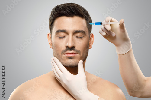 Young man isolated on gray background receiving botox injection from beauty doctor in sterile gloves in forehead to reduce aging, eyes closed