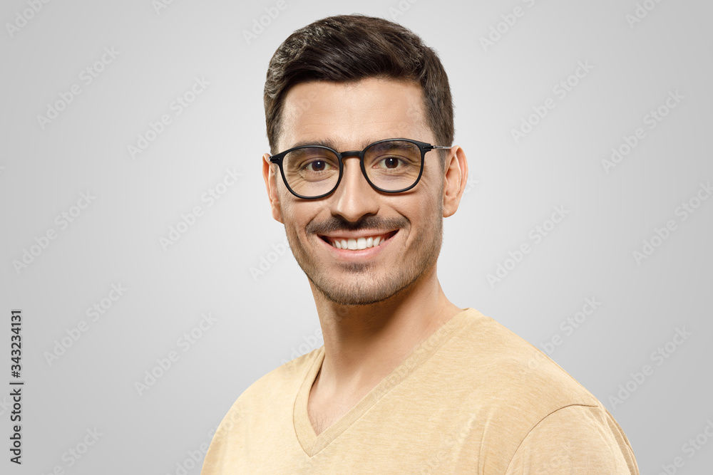 Close-up portrait of young man wearing beige t-shirt and trendy glasses, looking at camera with happy smile, isolated on gray background. Eyewear ads concept Close-up portrait of young man wearing beige t-shirt and trendy glasses, looking at camera with happy smile, isolated on gray background. Eyewear ads concept
