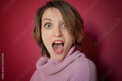 Close-up portrait of young emotional woman with short hair dressed in pink sweater is posing in the studio on bright pink background