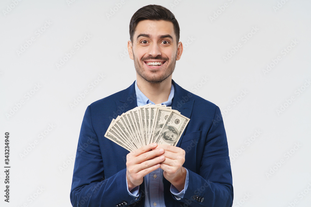 Young businessman feeling happy about getting increased income and receiving profit from new startup, holding dollars in cash in hands, smiling, isolated on gray background Young businessman feeling happy about getting increased income and receiving profit from new startup, holding dollars in cash in hands, smiling, isolated on gray background