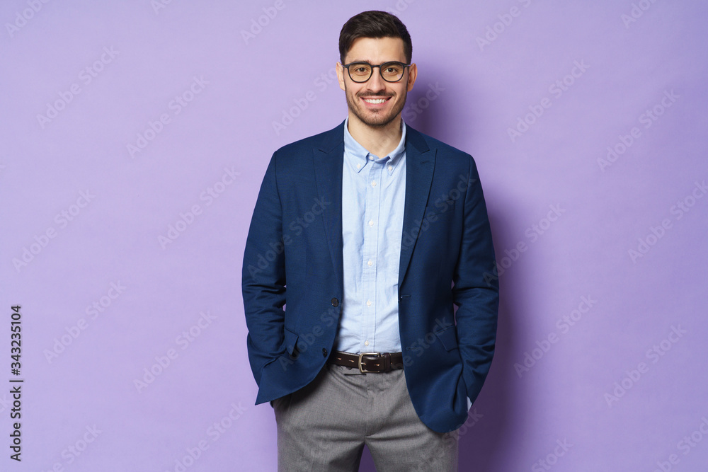 Young businessman, wearing glasses, standing isolated against purple background, feeling relaxed and confident, smiling positively, ready to help and support Young businessman, wearing glasses, standing isolated against purple background, feeling relaxed and confident, smiling positively, ready to help and support