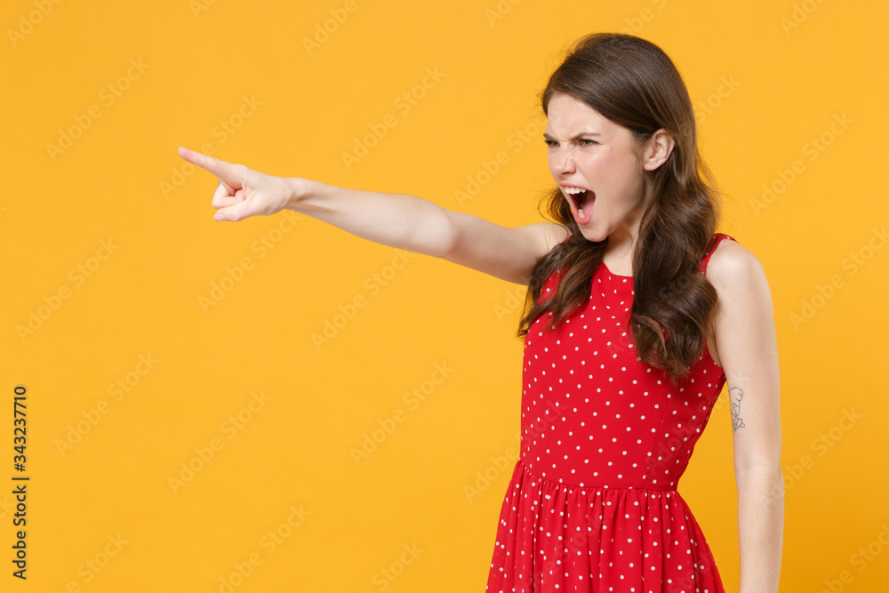 Irritated young brunette woman girl in red summer dress posing isolated on yellow wall background studio. People lifestyle concept. Mock up copy space. Pointing index finger aside, screaming swearing. Irritated young brunette woman girl in red summer dress posing isolated on yellow wall background studio. People lifestyle concept. Mock up copy space. Pointing index finger aside, screaming swearing.