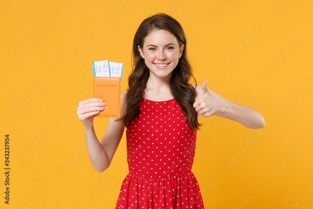 Smiling young brunette woman girl in red summer dress posing isolated on yellow background studio. People lifestyle concept. Mock up copy space. Hold passport tickets boarding pass, showing thumb up. Smiling young brunette woman girl in red summer dress posing isolated on yellow background studio. People lifestyle concept. Mock up copy space. Hold passport tickets boarding pass, showing thumb up.