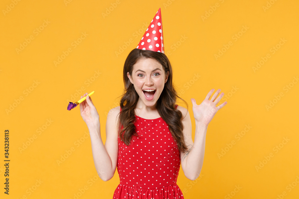 Excited young brunette woman girl in red summer dress, birthday hat posing isolated on yellow wall background studio portrait. Birthday holiday concept. Mock up copy space. Hold pipe, spreading hands.