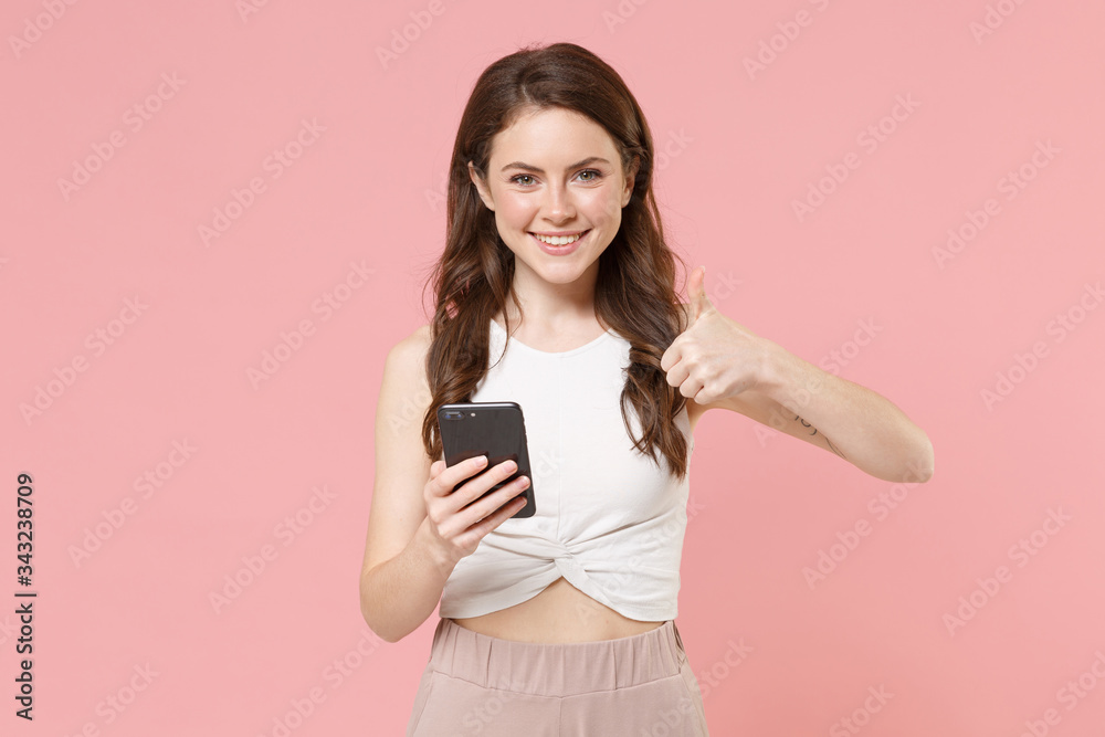 Smiling young brunette woman girl in light casual clothes isolated on pastel pink background. People lifestyle concept. Mock up copy space. Using mobile phone, typing sms message, showing thumb up. Smiling young brunette woman girl in light casual clothes isolated on pastel pink background. People lifestyle concept. Mock up copy space. Using mobile phone, typing sms message, showing thumb up.