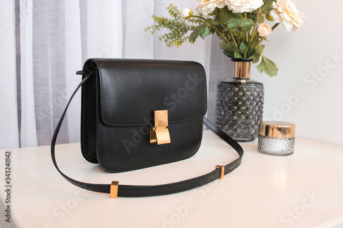 Black women’s leather bag on a white table with a vase, flowers and a clock in the background photo