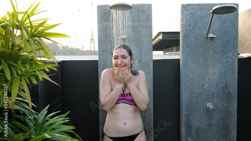 lady in bikini with long loose hair trembles under cold water pressure standing in hotel outdoor pool shower near tropic plant