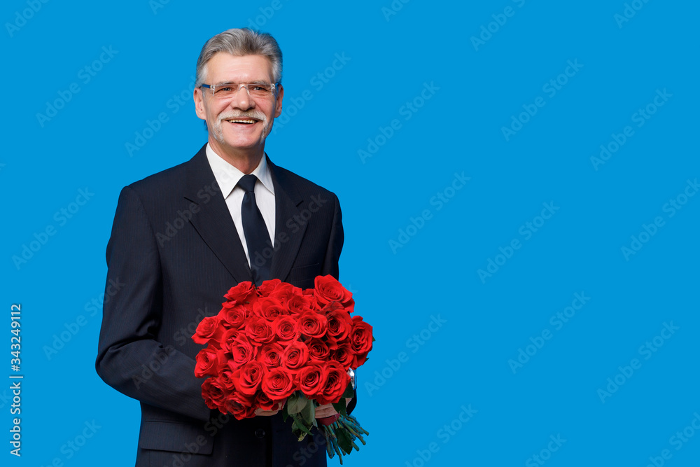 Older happy senior man with a bouquet of red rose flowers waiting on a date staying on blue background, copy space Older happy senior man with a bouquet of red rose flowers waiting on a date staying on blue background, copy space