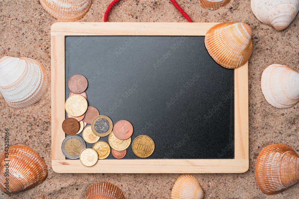 Blank blackboard with seashells and Euro coins on a golden beach sand. Summer travel expenses abstract concept. Copy space in the center.