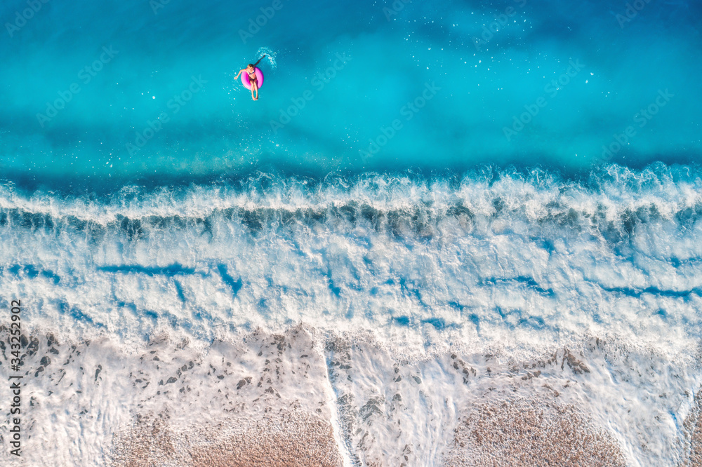 Aerial view of a young woman swimming with the donut swim ring in the clear blue sea with waves at sunset in summer. Tropical landscape with girl, azure water, sandy beach. View from above. Travel
