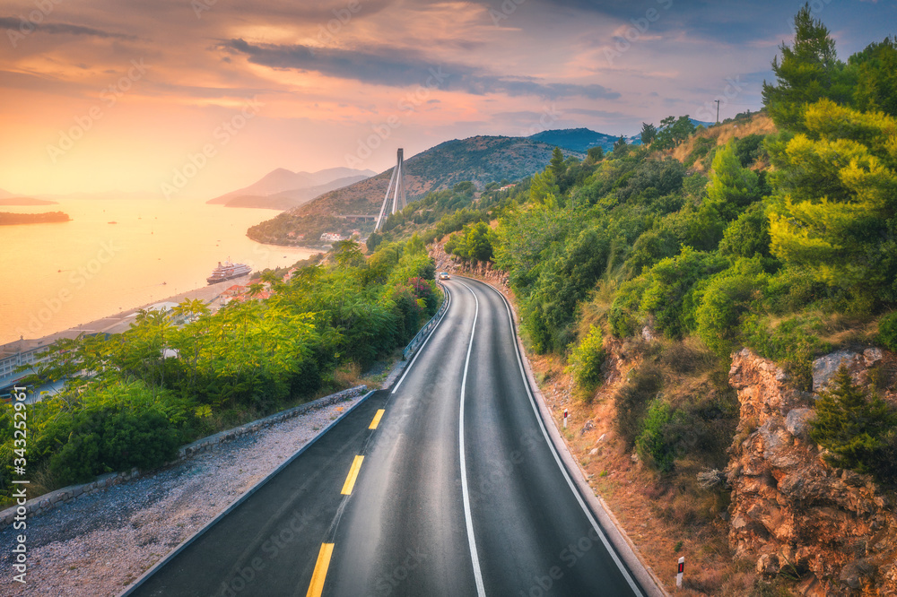 Obraz premium Aerial view of perfect mountain road and beautiful green forest at colorful sunset in summer. Dubrovnik, Croatia. Top view of road, sea, mountain, sky. Landscape with highway, sea coast, gold sunlight