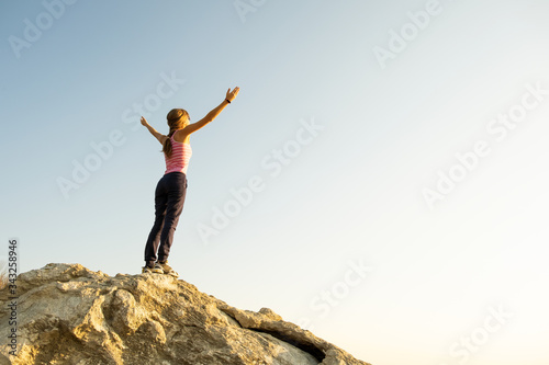 Young woman hiker standing alone on big stone in mountains. Female tourist raising her hands up on high rock in morning nature. Tourism  traveling and healthy lifestyle concept.