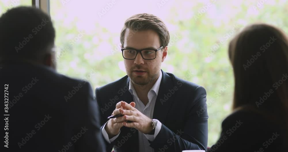 Successful young businessman in eyewear explaining business growth strategy to interested multiracial colleagues in office. Skilled male manager wearing suit, holding negotiations with coworkers.