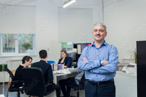 A portrait of a successful smiling senior businessman with his hands folded, his young team is on the background having a meeting in their office